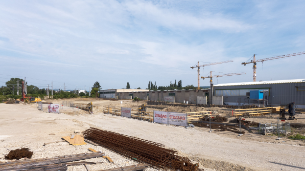 Baustelle mit verschiedenen Materialien, Geräten, Gebäuden, Kränen, Bäumen und einem bewölkten Himmel.