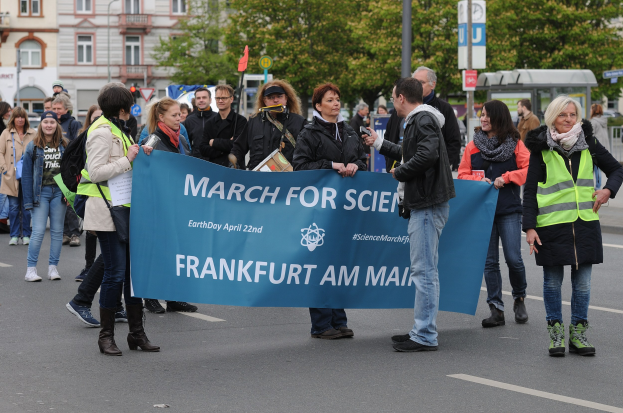 Gruppe von Menschen marschiert die Straße entlang und hält ein "March for Science Frankfurt am Main"-Schild, mit Bäumen, Gebäuden und einem klaren Himmel im Hintergrund.