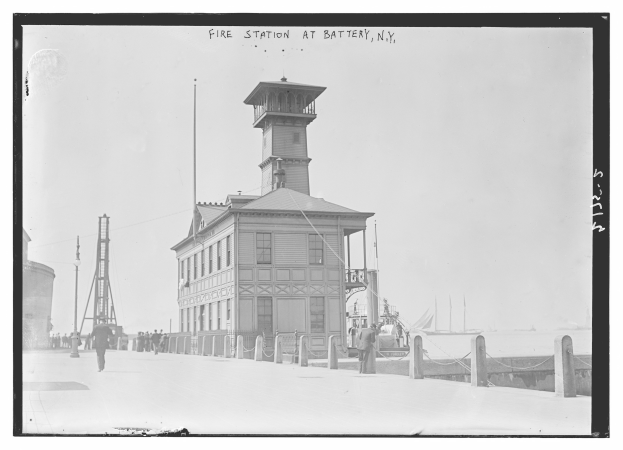 Schwarz-weiß-Foto einer Feuerwache an der Battery, New York, mit Menschen, Pfählen, einem Zaun, Booten auf dem Wasser und einem bewölkten Himmel.