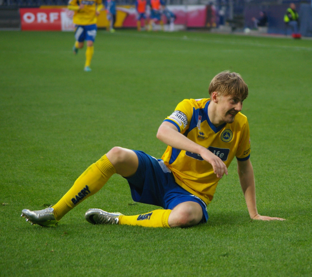 Ein Fußballer kniet auf dem Boden mit dem Kopf in den Händen, umgeben von Menschen in Sportkleidung, mit unscharfen Stadionelementen im Hintergrund.