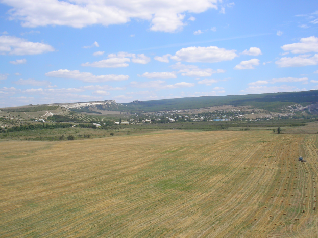 Ein Traktor, der auf einem kultivierten Feld umgeben von Bäumen, Gebäuden, Hügeln und einem bewölkten Himmel arbeitet.