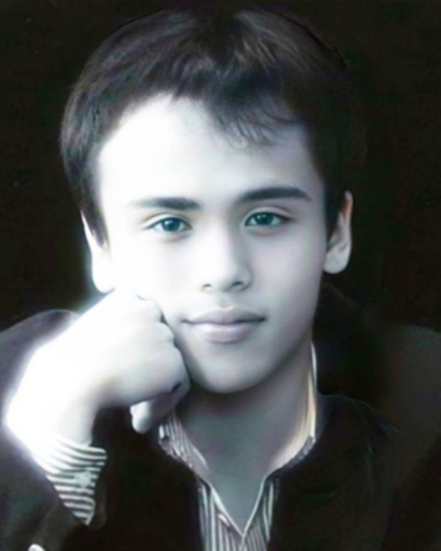 Ein junger Mann in einer schwarzen Jacke posiert selbstbewusst vor einem dunklen Hintergrund mit entschlossener Miene.