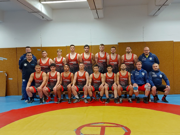 Eine Gruppe von Männern, einige sitzen auf Stühlen und andere stehen, posiert zusammen auf einer Ringer-matte mit einer Wand und Deckenlampen und Rohren im Hintergrund, was auf ein Ringerteam hinweist, das einen Sieg feiert.