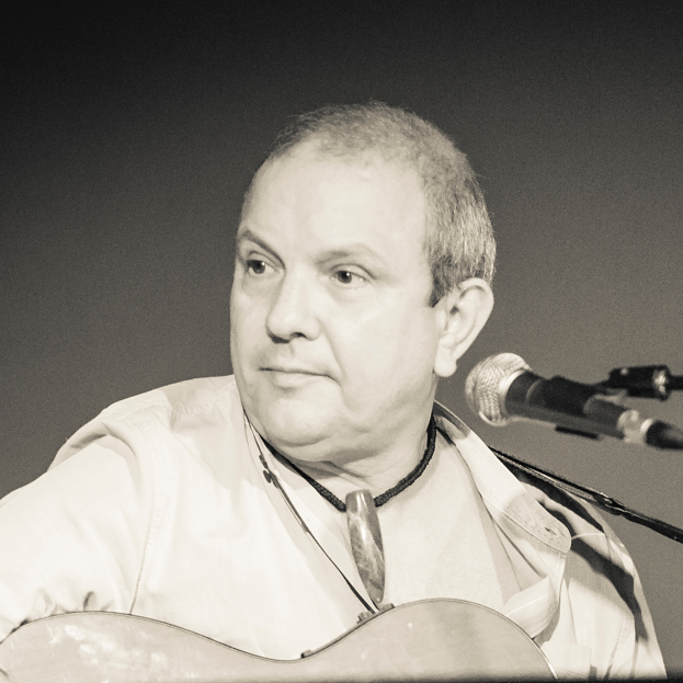 Ein Mann in einem weißen Hemd spielt eine Gitarre vor einem Mikrofon, mit einem ernsten Gesichtsausdruck, vor einem schwarz-weißen Hintergrund.
