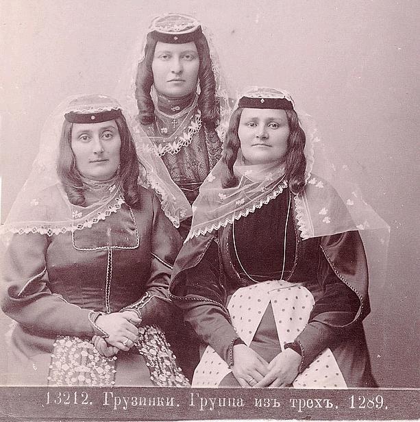 Drei Frauen in traditionellen russischen Hochzeitskleidern mit aufwendiger Stickerei und Perlen stehen vor einer weißen Wand in einem Schwarz-Weiß-Foto, das als russisches Hochzeitsfoto bezeichnet wird.