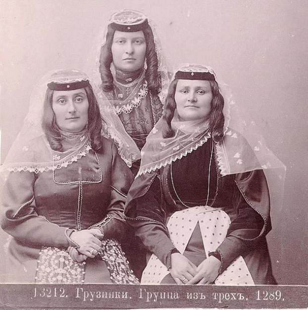 Drei Frauen in traditionellen russischen Hochzeitskleidern mit aufwendiger Stickerei und Perlen stehen vor einer weißen Wand in einem Schwarz-Weiß-Foto, das als russisches Hochzeitsfoto bezeichnet wird.