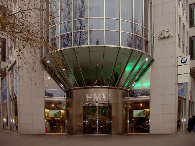 Außenansicht des BMW-Hauptquartiers in Frankfurt, Deutschland, mit einem modernen Glasgebäude mit Text darauf, Menschen vorneweg und einem Baum links.