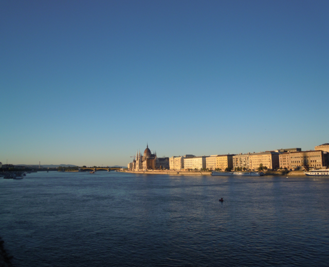 Luftaufnahme des Donauufers in Budapest, Ungarn, mit Booten auf dem Wasser, Gebäuden mit Fenstern, Bäumen, einer Brücke und einem Himmel im Hintergrund.