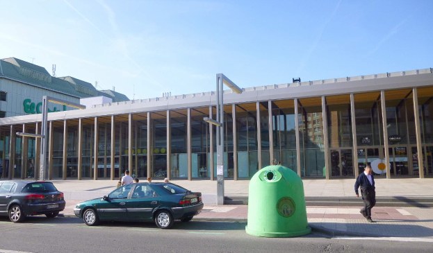 Grüne Mülltonne auf einer Stadtstraße neben einem gläsernen Gebäude mit Säulen, mit Fahrzeugen und Passanten im Blickfeld und einem bewölkten Himmel im Hintergrund.
