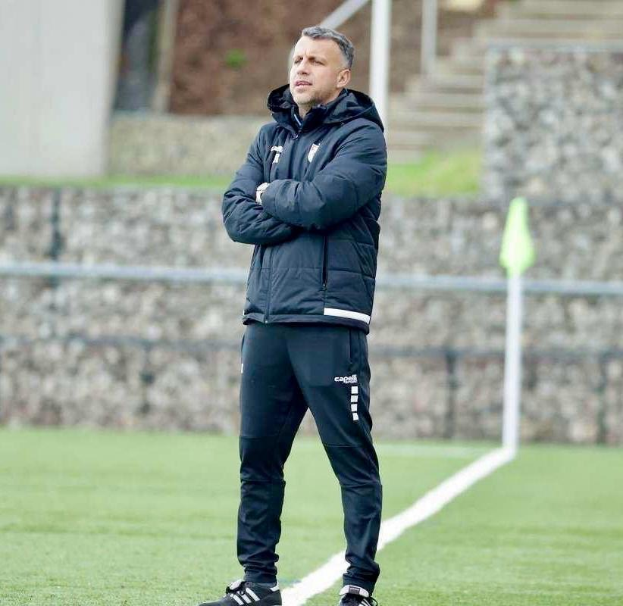 Ein Fußballtrainer steht auf einem Feld mit verschränkten Armen, trägt eine schwarze Jacke und Hose und befindet sich vor einem Zaun, Treppe und Wand während eines Trainingssitzungs.