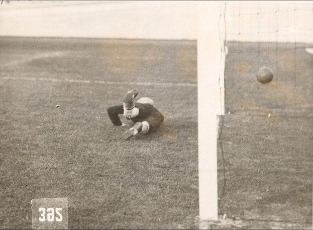 Schwarzes und weißes Foto eines Fußballspielers in der Mitte des Spiels, der einen Ball in der Nähe eines Torpfostens mit einem Netz auf der rechten Seite und Text unten fängt.