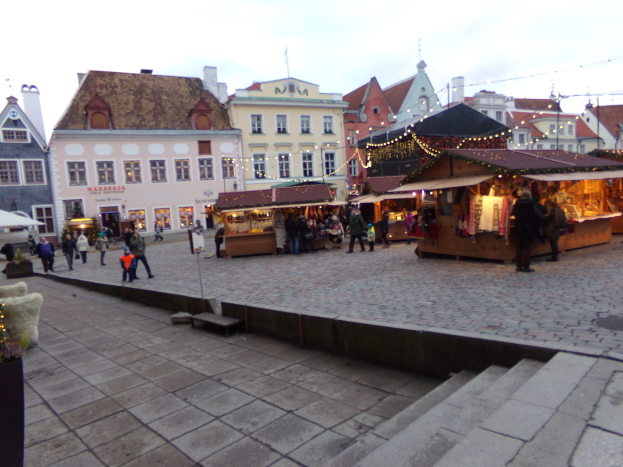 Ein lebendiger Weihnachtsmarkt in Tallinn, Estland, mit Menschen um beleuchtete und geschmückte Stände, Gebäuden mit Fenstern im Hintergrund, Treppen, die zum Markt führen, und Töpfen mit Pflanzen.