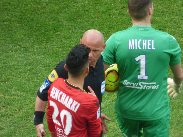 Schiedsrichter spricht mit zwei Fußballspielern auf einem grünen Feld, ein Spieler hält einen Ball.