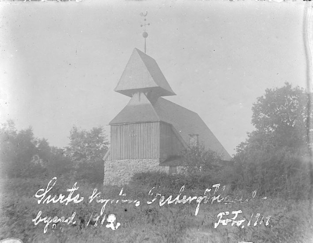 Schwarzes und weißes Foto einer Kirche mit einem Kirchturm, umgeben von Bäumen und Pflanzen unter einem bewölkten Himmel, beschriftet mit "Kirche des Heiligen Grabes" unten.