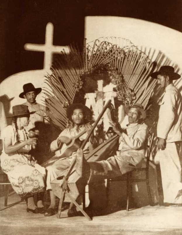 Schwarzes und weißes Foto einer Mariachi-Band, die vor einer Kirche mit einem Kreuz an der Wand dahinter sitzt, einige halten Instrumente und tragen Hüte.
