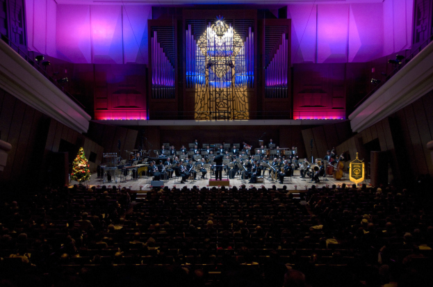 Großer Saal mit Publikum bei einem Konzert auf der Bühne, mit sitzenden Musikern und einem Weihnachtsbaum im Hintergrund mit geschmückten Wänden.