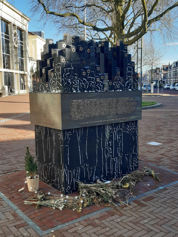 Denkmal in einem Stadtplatz für Holocaust-Opfer, mit einem Baum davor, einem Topf mit einer Pflanze auf dem Boden, verstreuten trockenen Blättern und im Hintergrund Gebäude, Fahrzeuge, Masten und Bäume.