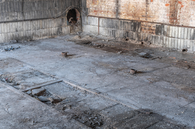 Ein leerer Raum mit einer Backsteinwand mit einem Loch und einem Boden, der mit verschiedenen Gegenstönden bedeckt ist, was auf ein verlassenen Geböude hindeutet.