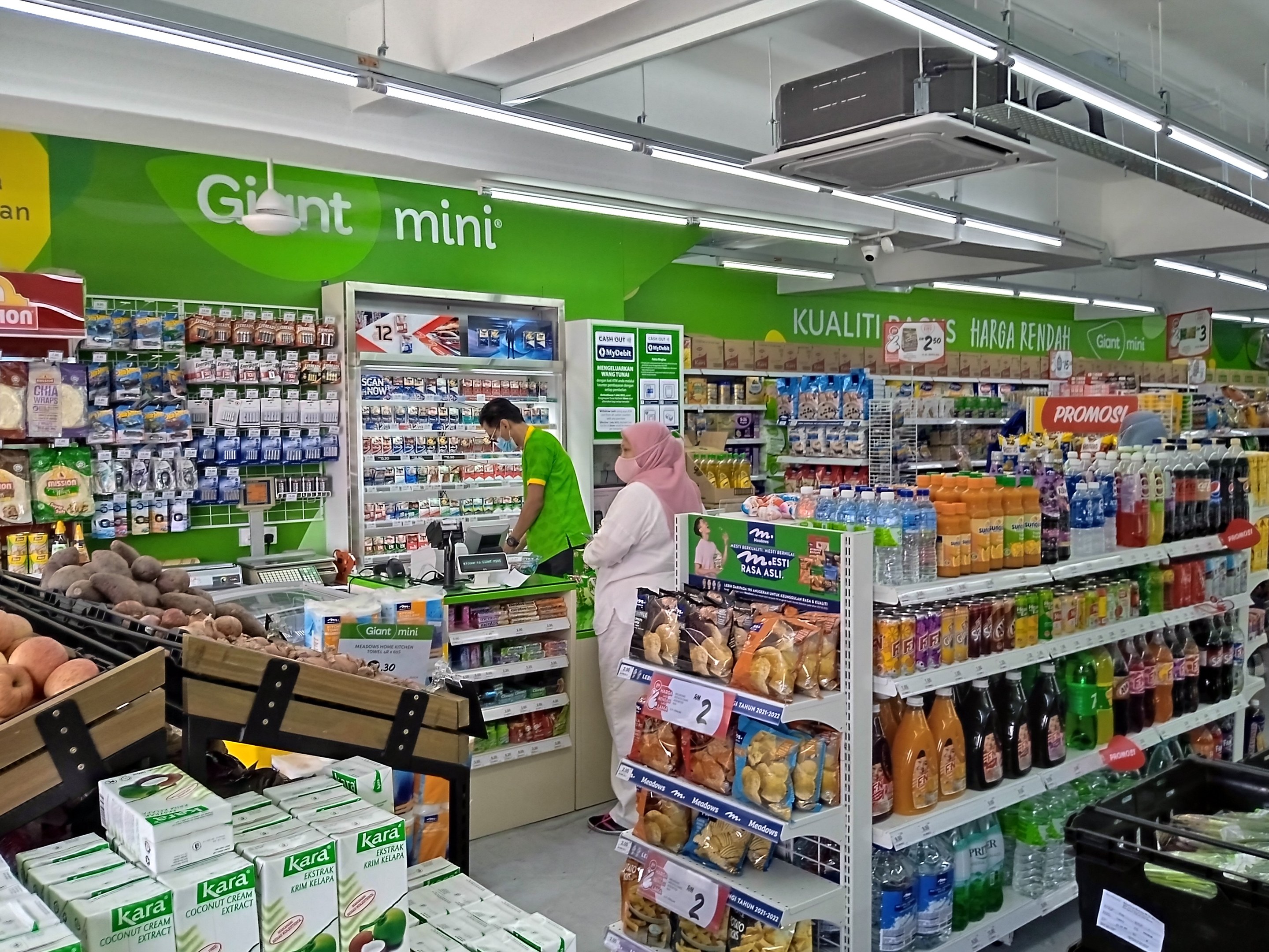 Ein Geschäft mit Regalen voller verschiedener Lebensmittel wie Flaschen, Schachteln und Früchte sowie Deckenbretter, Lampen und ein Projektor, was auf einen neu eröffneten Mini-Supermarkt in Malaysia hinweist.