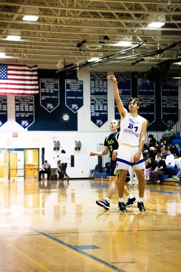 Ein Mann in Sportschuhen wirft einen Basketball in einer Turnhalle, umgeben von Zuschauern auf Tribünen und Bannern und einer Fahne an der Wand im Hintergrund.