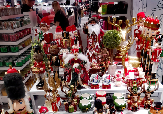 Ladeninnenraum mit Weihnachtsdekorationen einschließlich Spielzeug, Tassen und anderen Gegenständen auf Tischen und Regalen, mit Menschen, Lichtern und einer Wand im Hintergrund.