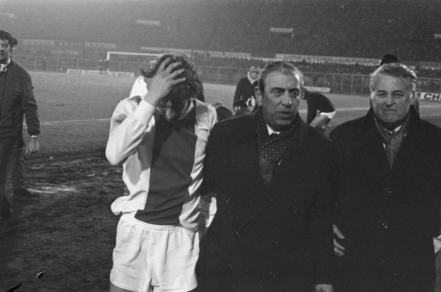 Schwarzes und weißes Bild einer Gruppe von Männern auf einem Fußballfeld mit einem Stadion im Hintergrund, die nach einem Spiel bedrückt aussehen.
