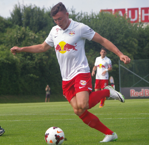 Ein Mann in einem Fußballtrikot kickt einen Ball auf einem grünen Feld mit Bäumen und einem klaren blauen Himmel im Hintergrund, mit einer Tafel mit der Aufschrift "RB Leipzig" sichtbar.
