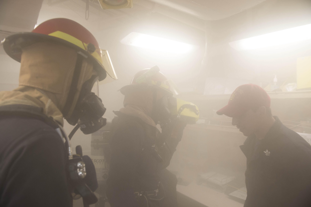 Feuerwehrleute mit Helmen und Rauch aus dem Mund, einer hält eine Kamera, während einer Übung mit Hintergrundbeleuchtung und -gegenständen.