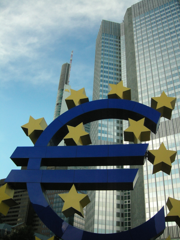 Euro-Zeichen vor dem Hauptquartier der Europäischen Zentralbank in Frankfurt, Deutschland, mit dem Gebäude umgeben von Bäumen unter einem bewölkten Himmel.