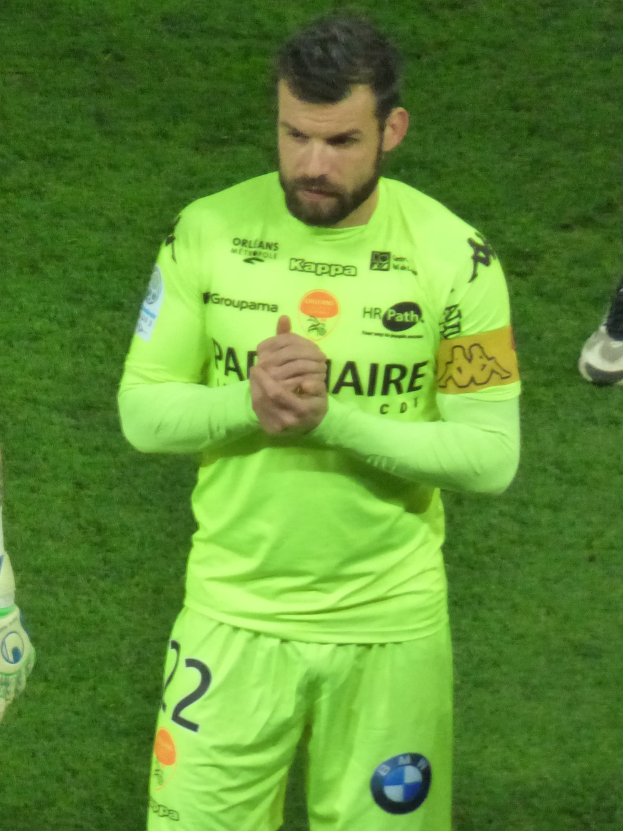 Ein Torwart in einem gelben Trikot klatscht auf einem Fußballfeld in die Hände, trägt ein grünes Hemd und Shorts und hat ein weiteres Bein links neben sich.