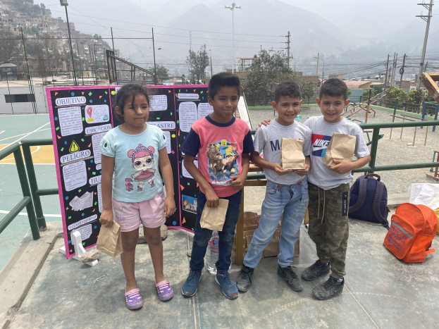 Gruppe von Kindern auf einem Basketballfeld mit Papiertüten, Tüten und Gegenständen rechts und einer Tafel mit Text links, vor einem Hintergrund aus Zäunen, Pfosten, Straßenlaternen, Bäumen, Gebäuden, Hügeln und Himmel.