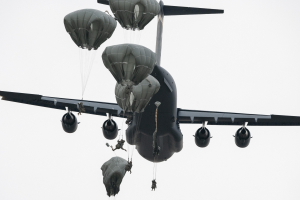 Ein großes Militärflugzeug mit mehreren Fallschirmen, das gegen einen klaren blauen Himmel fliegt.