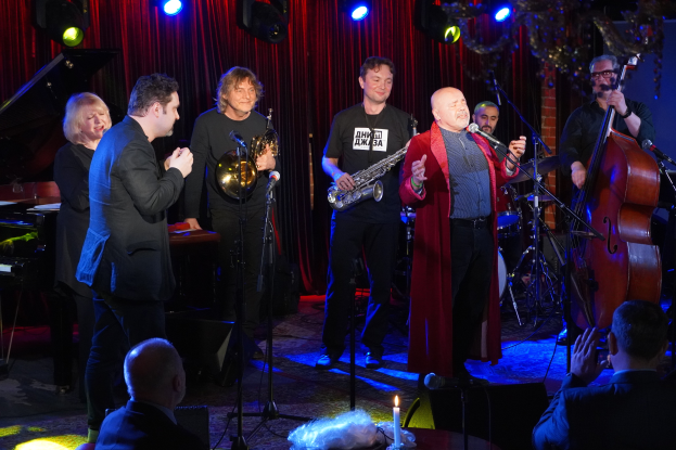 Eine Gruppe von Menschen auf einer Bühne mit Musikinstrumenten, Mikrofonen und einer Kerze, die vor einem sitzenden Publikum spielt, unter Scheinwerfern und einem Vorhang.