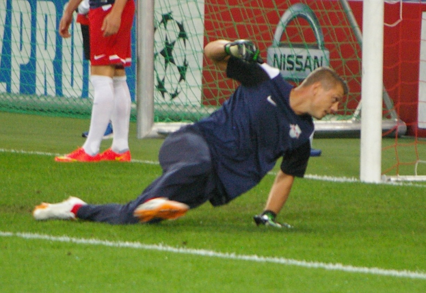 Fußballtorwart taucht ab, um einen Ball vor einem Tor mit zwei Personen im Hintergrund, einem Netz und einer Banner mit Text zu fangen.