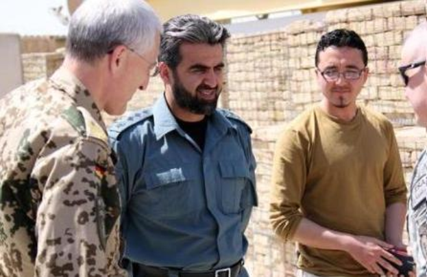 Eine Gruppe von Männern steht vor einer Backsteinwand, einer hält ein Objekt, während der Mann in der Mitte von der irakischen Armee interviewt wird, was darauf hindeutet, dass er der US-Verteidigungsminister ist.