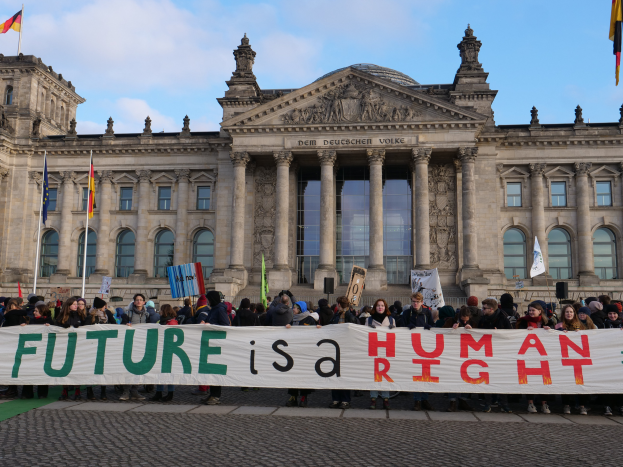 Eine Gruppe von Menschen hält ein Banner mit der Aufschrift "Zukunft ist ein Menschenrecht" vor dem Reichstagsgebäude in Berlin, Deutschland, das mit Fenstern, Säulen, Bögen und umgeben von Fahnenmasten geschmückt ist.