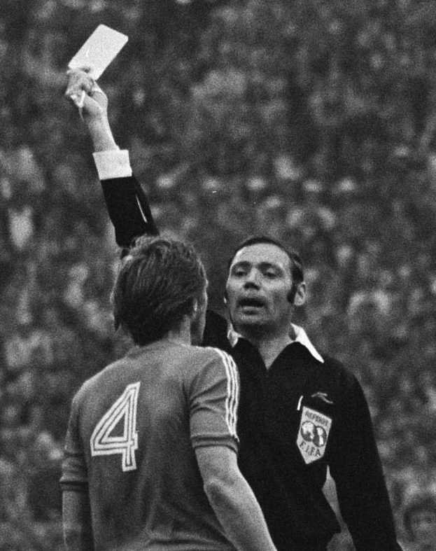 Ein Schiedsrichter in einem Schwarz-Weiß-Foto hält eine weiße Karte einem Fußballspieler hin, wobei der Hintergrund unscharf ist.