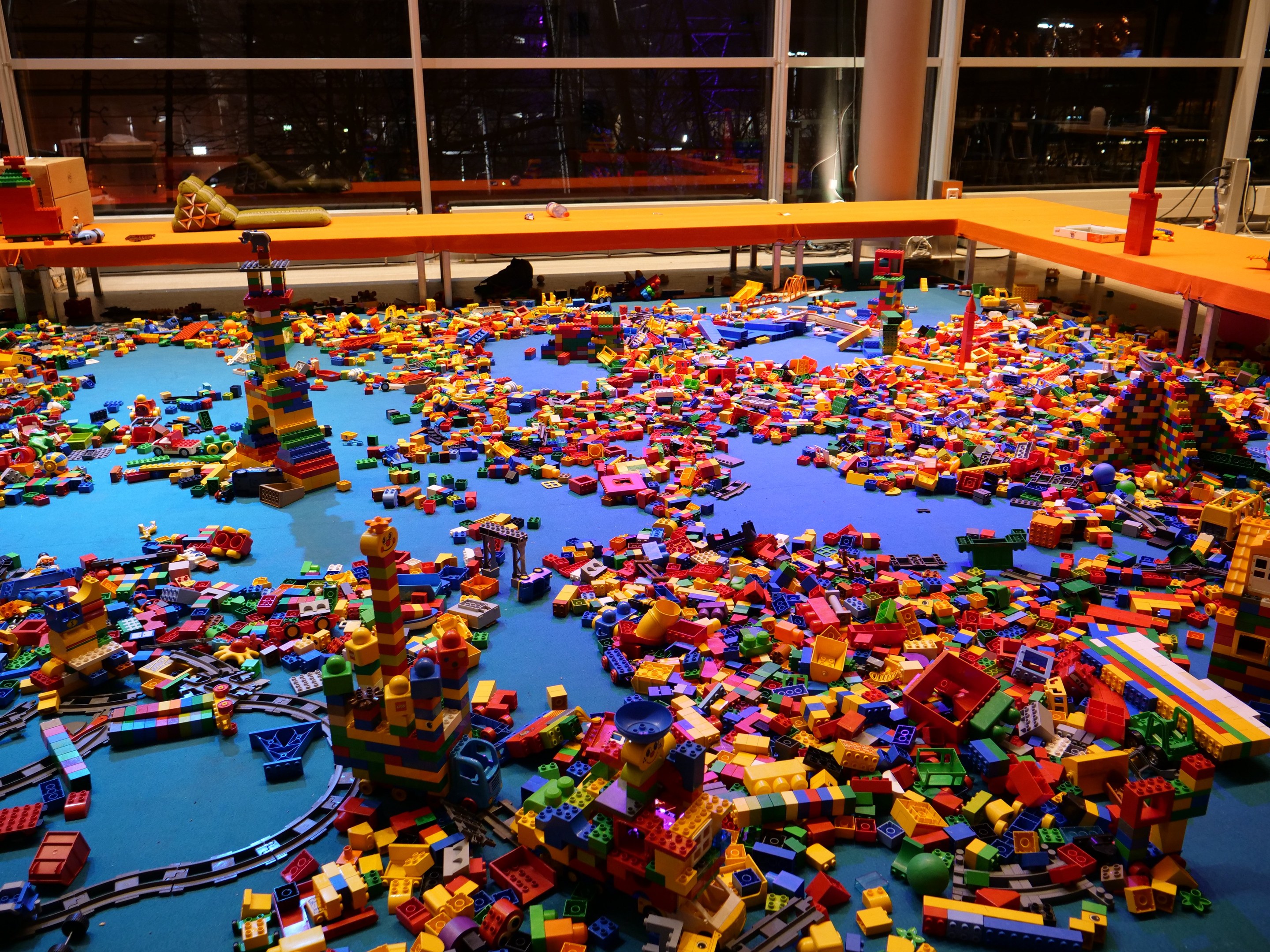 Ein großer Raum mit farbigen Lego-Bausteinen, die auf dem Boden verstreut sind, umgeben von Tischen und Säulen, mit Glasfenstern im Hintergrund, die Menschen und Lichter zeigen.