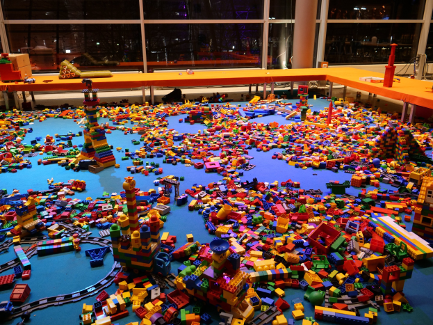 Ein großer Raum mit farbigen Lego-Bausteinen, die auf dem Boden verstreut sind, umgeben von Tischen und Säulen, mit Glasfenstern im Hintergrund, die Menschen und Lichter zeigen.