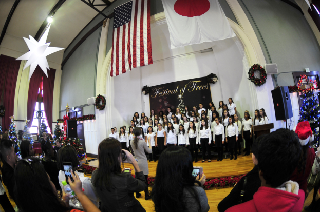 Eine Gruppe von Menschen auf einer Bühne vor einem Publikum, mit Weihnachtsdekoration, einem Rednerpult, Lautsprechern, einer Texttafel, Vorhängen, Fahnen und Deckenleuchten.