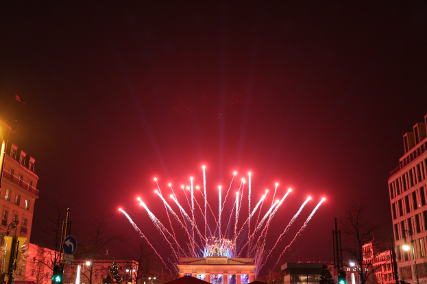 Das Bild zeigt eine nächtliche Straßenansicht in Berlin mit dem Brandenburger Tor in der Mitte, umgeben von beleuchteten Gebäuden, Bäumen, Pfosten, Verkehrszeichen, Schildern und Fahnen, mit Feuerwerk am Himmel über dem Tor.