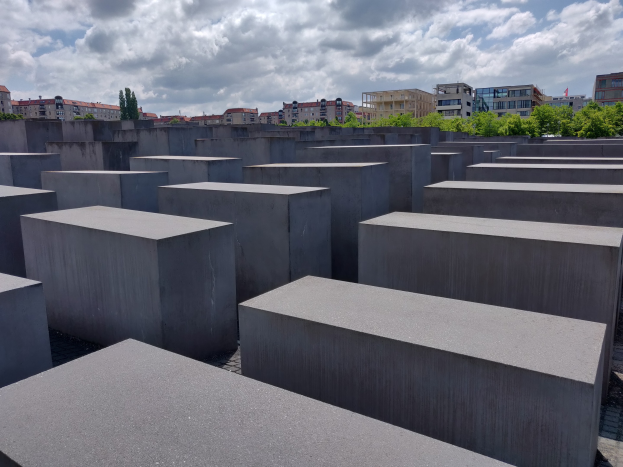 Konkreter Holocaust-Mahnmal mit rechteckigen Blöcken in einem Raster angeordnet, umgeben von Bäumen, Gebäuden und einer bewölkten Himmel.