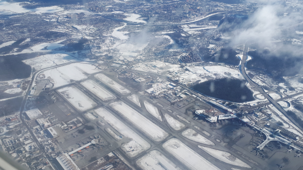 Luftaufnahme eines schneebedeckten Flughafens mit Gebäuden, Straßen, Brücken, Wasser, Bäumen und Wolken am Himmel.