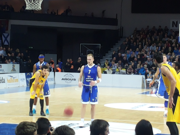 Eine Gruppe von Männern in blauen und gelben Uniformen spielt Basketball auf einem Court mit Zuschauern in den Rängen, während eines Spiels zwischen Fenerbahçe und Real Madrid in der 7days Eurocup Regular Season Round 2.