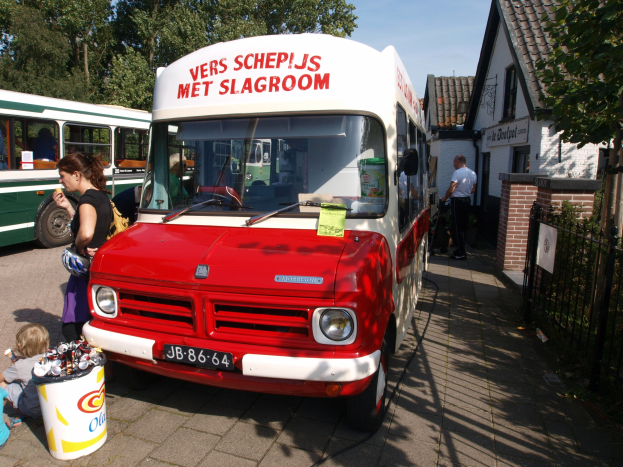 Roter und weißer Eiscreme-Wagen auf einer Straße mit Menschen in der Nähe und einem Eimer Eiscreme davor, vor einem Hintergrund aus Gebäuden, Bäumen und einem klaren blauen Himmel.