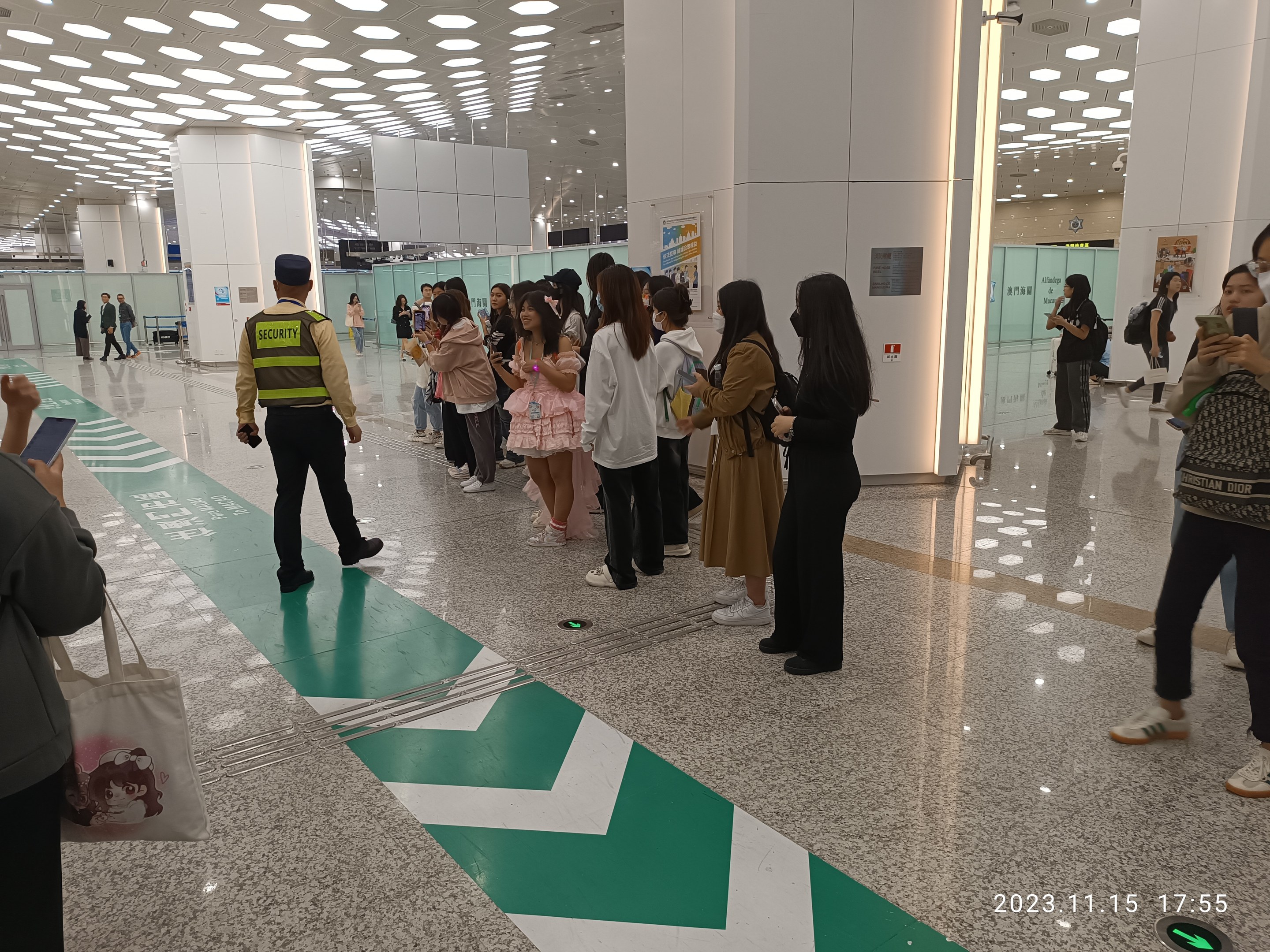 Menschen in einer Reihe an einem Flughafen-Sicherheits-Check-in, einige halten Taschen und Mobiltelefone, mit Säulen und Deckenleuchten im Hintergrund.