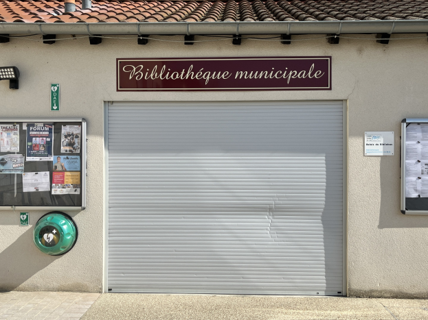 Außenansicht eines Gebäudes mit der Aufschrift "Bibliothèque municipale" mit einem Rollladen, Plakaten und einem beleuchteten Weg zur Eingangstür.