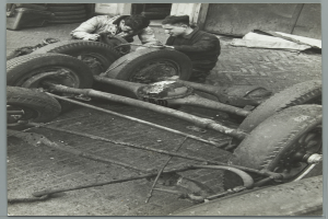 Zwei Männer in einer Garage, die Werkzeuge halten, arbeiten an einem Auto umgeben von Reifen und anderen Gegenständen, dargestellt in Schwarz-Weiß.