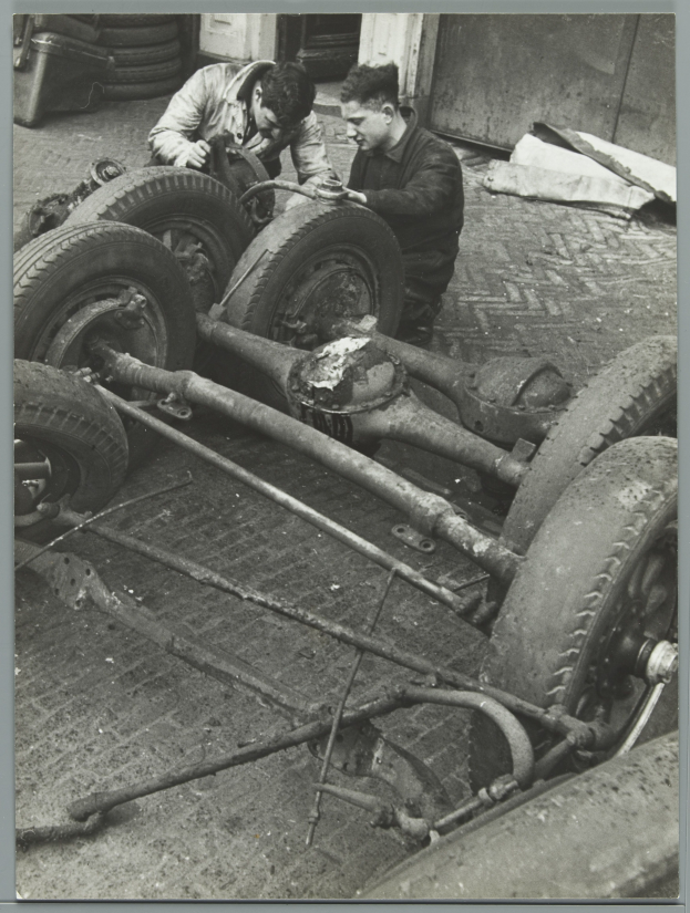 Zwei Männer in einer Garage, die Werkzeuge halten, arbeiten an einem Auto umgeben von Reifen und anderen Gegenständen, dargestellt in Schwarz-Weiß.