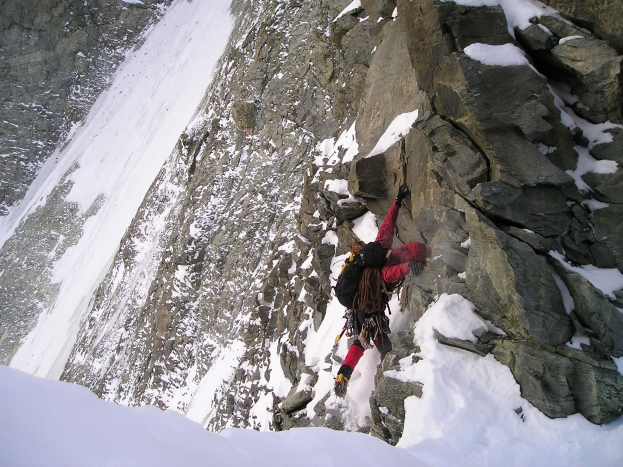 Eine Person in einer roten Jacke und mit einem Rucksack klettert einen steilen, schneebedeckten Berghang hinauf.